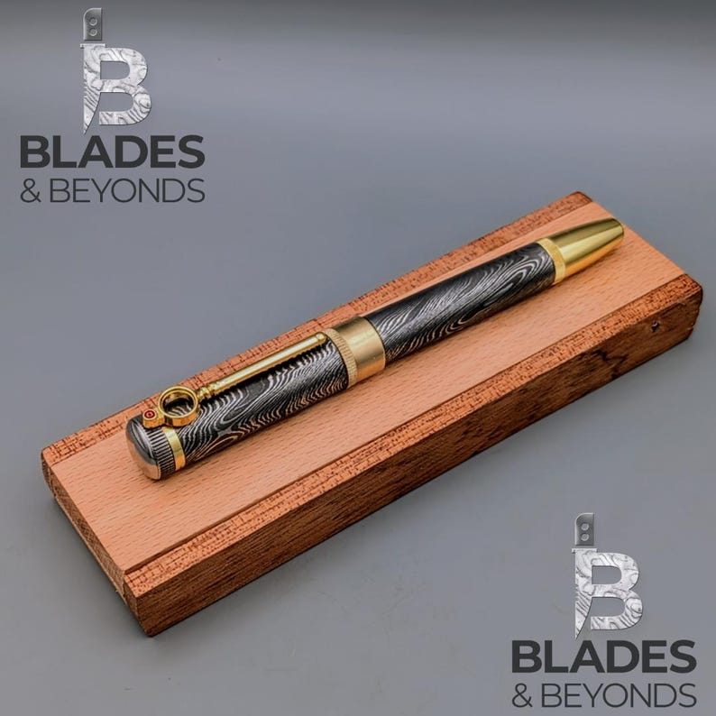 May include: A Damascus steel pen with gold accents rests on a wooden base. The pen features a patterned design in black and silver, with a gold-colored tip and trim. The pen is displayed on a rectangular, reddish-brown wooden base. The logo "Blades & Beyonds" is visible.