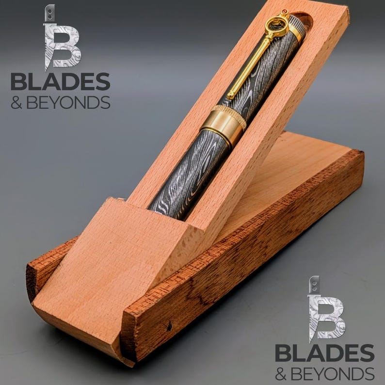 May include: A Damascus steel pen with gold accents rests in an open wooden box. The pen features a key-shaped clip and a patterned barrel. The box is made of light-colored wood and is angled for display. The logo "Blades & Beyonds" is visible.