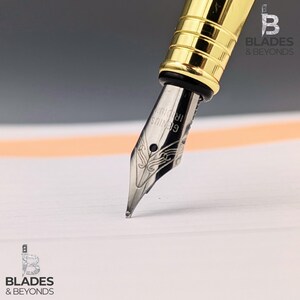 May include: Close-up of a gold and silver fountain pen with the nib touching a white paper. The pen has a gold barrel and a silver nib with the text "GENIUS IRIDIUM". The pen is angled towards the bottom right corner.