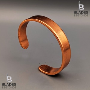 Puede incluir: Una pulsera de puño de cobre con un diseño sencillo y abierto. La pulsera tiene un perfil plano y rectangular y un color marrón rojizo cálido. El metal tiene una superficie ligeramente texturizada. El logotipo "Blades & Beyonds" está en las esquinas superior derecha e inferior izquierda.