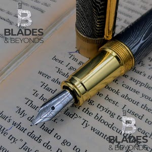 May include: A close-up of a luxury fountain pen with a gold-colored barrel and nib. The pen's cap and barrel feature a black and silver woodgrain pattern. The pen is resting on a book with printed text. The logo "BLADES & BEYONDS" is visible.