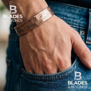 May include: Copper cuff bracelet with a unique, swirling pattern. The bracelet is worn on a wrist, with the hand in a blue jeans pocket. The logo "Blades & Beyonds" is visible in the image.