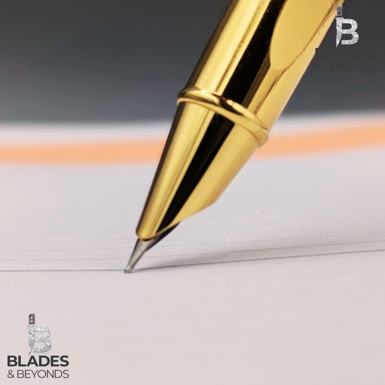 Puede incluir: Primer plano de un bol&iacute;grafo dorado escribiendo sobre papel blanco. El bol&iacute;grafo tiene un acabado met&aacute;lico brillante y est&aacute; inclinado hacia abajo, con la punta tocando el papel. El fondo est&aacute; borroso, con un toque de naranja y gris.