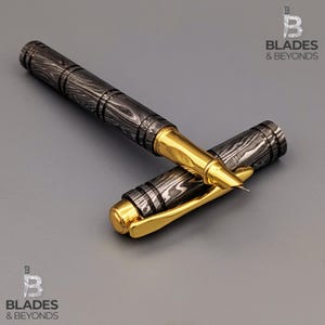 Puede incluir: Un bol&iacute;grafo roller con un barril gris oscuro estampado y detalles dorados. La tapa del bol&iacute;grafo est&aacute; quitada, revelando la punta dorada y el dise&ntilde;o del bol&iacute;grafo. El bol&iacute;grafo est&aacute; sobre una superficie gris. El logotipo "Blades & Beyonds" est&aacute; en las esquinas superiores derecha e inferior izquierda.