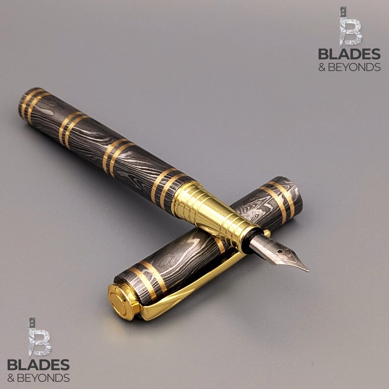 May include: A fountain pen with a black and gold patterned barrel and gold accents. The pen's cap is off, revealing the nib. The pen is lying on a gray surface.