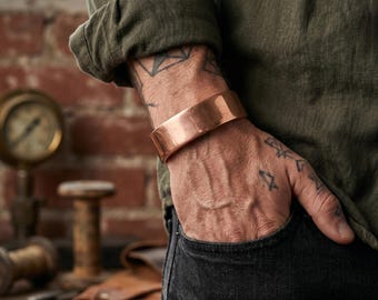 Könnte beinhalten: Ein breites, poliertes Kupfer-Armband mit einer glatten, reflektierenden Oberfläche und einem einfachen, offenen Design. Das Metallband hat einen warmen, metallisch rötlich-braunen Farbton und liegt fest am Handgelenk an, was eine klare Ästhetik zeigt.