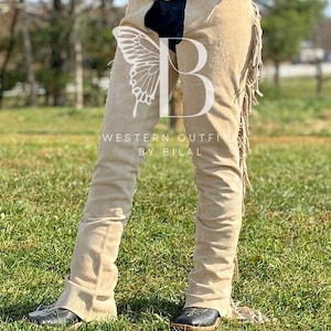 Op de afbeelding: Beige suède chaps met franjes en een vlinderontwerp. De chaps worden over jeans gedragen en hebben een riem met een gesp. De tekst "WESTERN OUTFITS BY BILAL" is zichtbaar op de chaps.