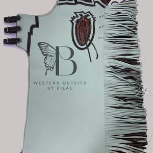 May include: Light blue leather chaps with brown leather detailing and fringe. The chaps have a butterfly graphic and the text "WESTERN OUTFITS BY BILAL". Decorative elements include a shield design and embossed leather.