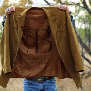 May include: A brown suede jacket with a brown satin lining. The lining features a butterfly design and the text "Western Outfits by Bilal".