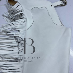 Puede incluir: Chaps de cuero blanco con flecos largos. Los chaps presentan un diseño calado decorativo y un cierre de hebilla plateada. El texto "WESTERN OUTFITS BY BILAL" está impreso en los chaps.