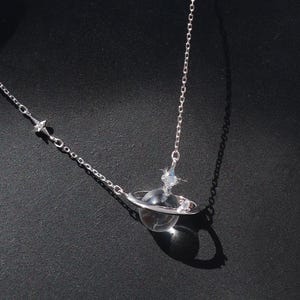 Peut inclure: Un délicat collier argenté avec un pendentif planète, un centre sphérique transparent et un anneau. La chaîne fine est ornée de petites étoiles. Le pendentif est présenté sur un mannequin noir.