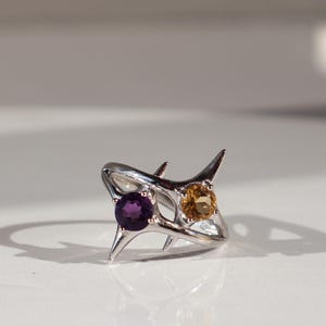 May include: A silver ring featuring a unique, spiky design. The ring is adorned with two gemstones: a round, deep purple amethyst and a round, golden citrine. The ring is set against a white background, with shadows adding depth.
