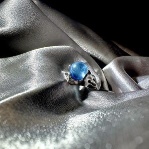 Op de afbeelding: Een zilveren ring met een grote, ronde, blauwe edelsteen. De band van de ring heeft een decoratief, opengewerkt ontwerp. De ring wordt gepresenteerd op een zilveren, zijdeachtige stof, waardoor een contrast ontstaat tussen de koele tinten van het metaal en de edelsteen.
