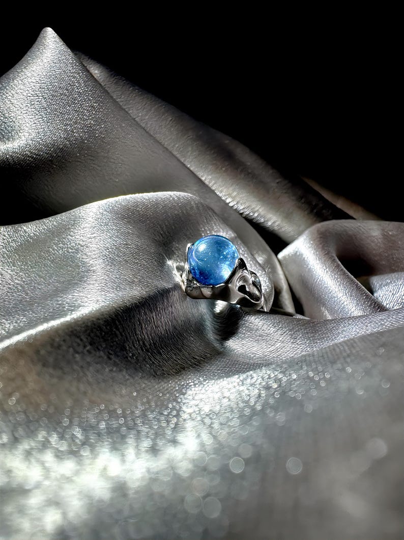 Op de afbeelding: Een zilveren ring met een grote, ronde, blauwe edelsteen. De band van de ring heeft een decoratief, opengewerkt ontwerp. De ring wordt gepresenteerd op een zilveren, zijdeachtige stof, waardoor een contrast ontstaat tussen de koele tinten van het metaal en de edelsteen.