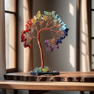May include: A copper wire tree sculpture adorned with colourful gemstones, representing the seven chakras. The tree stands on a dark grey stone base and is displayed on a wooden table.