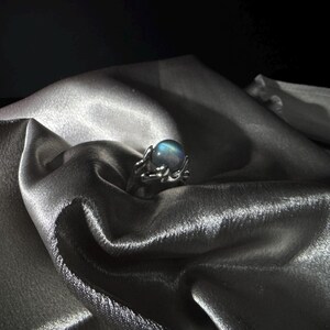 Op de afbeelding: Een zilveren ring met een grote, iriserende, ronde edelsteen. De ring heeft een delicate, takachtige vormgeving. De ring is geplaatst tegen een achtergrond van gedrapeerde, glinsterende, zilveren stof.