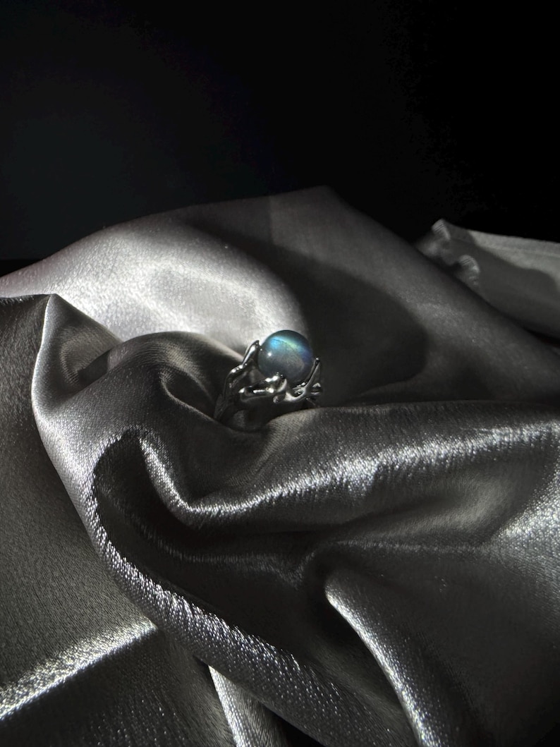 Op de afbeelding: Een zilveren ring met een grote, iriserende, ronde edelsteen. De ring heeft een delicate, takachtige vormgeving. De ring is geplaatst tegen een achtergrond van gedrapeerde, glinsterende, zilveren stof.
