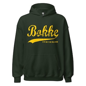 Puede incluir: Sudadera con capucha verde oscuro con capucha y bolsillo delantero. La sudadera presenta la palabra "Bokke" en texto amarillo estilizado, con la frase "IT'S IN THE BLOOD" debajo en texto amarillo más pequeño. La sudadera está hecha de un material suave.
