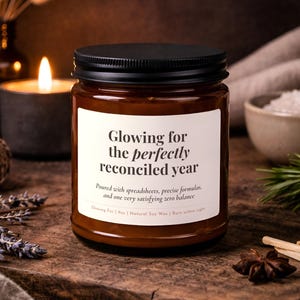 以下が含まれることがあります： 黒い蓋が付いた茶色のガラス製キャンドルジャー。白いラベルには「Glowing for the perfectly reconciled year. Poured with spreadsheets, precise formulas, and one very satisfying zero balance. Glowing For | 255g | Natural Soy Wax | Burn within sight.」と記載されています。
