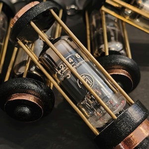 Può includere: Primo piano di diversi portachiavi in stile steampunk. Ogni portachiavi presenta un tubo di vetro trasparente racchiuso in una gabbia nera e color rame con aste dorate. Il tubo contiene componenti interni e marcature. I portachiavi sono su una superficie scura.