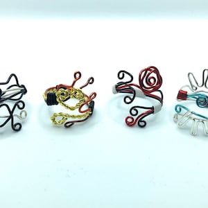 May include: Four wire rings with different designs. The first ring is black with a purple accent, the second is gold and red, the third is red and black with a white accent, and the fourth is silver with a turquoise accent.