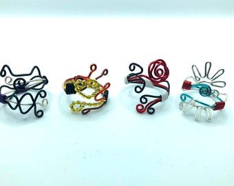 May include: Four wire rings with different designs. The first ring is black with a purple accent, the second is gold and red, the third is red and black with a white accent, and the fourth is silver with a turquoise accent.