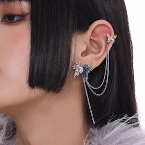 May include: A silver ear cuff and earring set featuring butterfly and floral designs. The ear cuff is attached to the earring with delicate chains. The earring has a blue and silver floral design. The brand name "SUMIYAKI" is visible at the bottom.