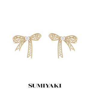 May include: A pair of gold-toned stud earrings shaped like bows with a delicate filigree pattern and a single clear round gemstone set in the center of each bow. The text SUMIYAKI is printed in black at the bottom of the image.
