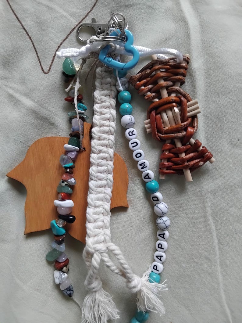 May include: A selection of handmade keyrings. One keyring has a macrame design, another features a beaded strand with the word "AMOUR PAPA", and a third is made of woven wood. The keyrings are attached to a silver clasp and a blue heart-shaped ring.