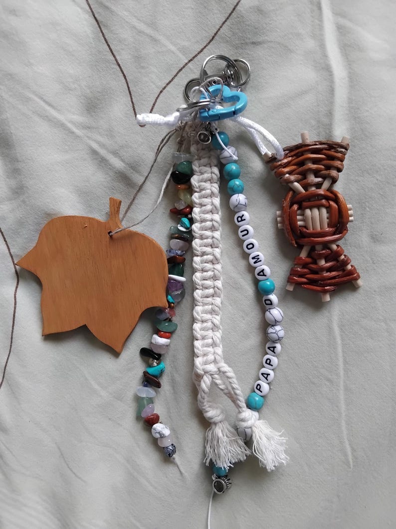 May include: A selection of handmade keyrings and charms. Features a wooden leaf, a woven basket, and beaded strands with turquoise, white, and colourful stones. One keyring spells "PAPA AMOUR" with white letter beads and turquoise accents.