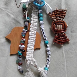 May include: A selection of handmade keyrings. One keyring has a macrame design with a tassel, another features a beaded strand with the words "AMOUR" and "PAPA", and a third is made of woven wood. Various colours of beads and stones are used.