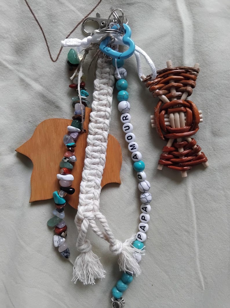 May include: A selection of handmade keyrings. One keyring has a macrame design with a tassel, another features a beaded strand with the words "AMOUR" and "PAPA", and a third is made of woven wood. Various colours of beads and stones are used.