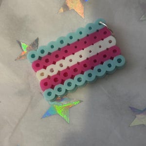 Puede incluir: Un llavero artesanal con la bandera del orgullo transgénero, hecho con cuentas de plástico de colores. El diseño de la bandera incluye rayas celeste, rosa, blanco, rosa y celeste. El llavero está rodeado de estrellas holográficas.