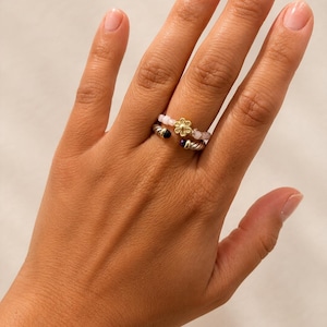 May include: A stacked ring set consisting of one beaded ring with light pink beads and a central gold-toned flower charm, and one silver-toned twisted cable ring featuring small dark purple stones at each end of the open band design.