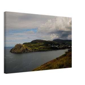 Puede incluir: Una vista panorámica de una costa rocosa con un pequeño pueblo enclavado en las colinas. El cielo está nublado con un toque de azul que se asoma. El agua está tranquila y refleja el cielo.