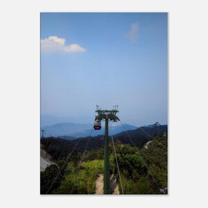 May include: A green cable car system with a red cable car traveling up a mountainside. The cable car is surrounded by lush green trees and a blue sky with white clouds.