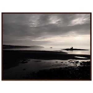 May include: A framed landscape photograph of a beach at dusk. The sky is overcast with dark clouds, and the water reflects the dim light. A small island with a castle silhouette is visible in the distance.