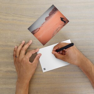 Op de afbeelding: Een persoon schrijft met een zwarte pen op een blanco witte ansichtkaart. Een tweede ansichtkaart toont een landschap met een silhouet van een persoon tegen een rood-oranje woestijn. De scène speelt zich af op een lichtbruin houten oppervlak.