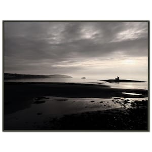 May include: A framed black and white photograph of a coastal scene under a cloudy sky. The foreground shows a dark beach with reflections. In the distance, a small island with a structure is visible, along with a boat on the water.