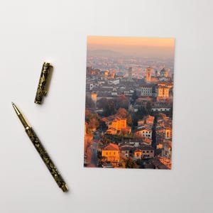 May include: A postcard featuring an aerial view of a European city at sunset, with buildings and towers bathed in warm light. A black and gold pen with floral detailing lies beside the postcard.