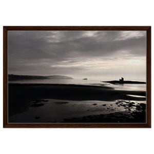 May include: A framed black and white landscape print. The image depicts a beach scene with a castle on a small island, a boat on the water, and a cloudy sky. The frame is dark brown.