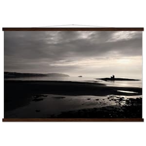 May include: A framed landscape print depicting a coastal scene under a cloudy sky. The image shows a dark beach, calm water, and a distant island with a structure. The print is mounted with wooden bars at the top and bottom.