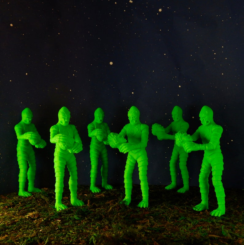 Pu&ograve; includere: Sei figure di plastica verde di una creatura della laguna nera, in piedi su una superficie verde ricoperta di muschio su uno sfondo blu scuro con stelle bianche.