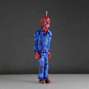May include: A red and blue zombie figurine with a blue jumpsuit and a red face. The figurine has a small antenna on its head and is standing on a white surface.