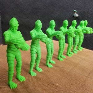 Pu&ograve; includere: Una fila di sette figurine di plastica verde di una creatura della laguna nera. Le figurine sono tutte nella stessa posa, con le braccia alzate e le mani giunte.