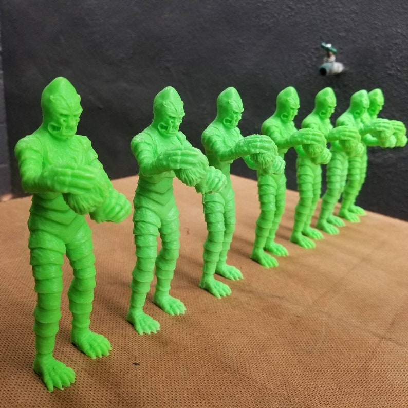 Pu&ograve; includere: Una fila di sette figurine di plastica verde di una creatura della laguna nera. Le figurine sono tutte nella stessa posa, con le braccia alzate e le mani giunte.