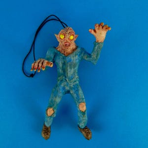 May include: A blue and brown monster figurine with glowing yellow eyes. The monster is wearing a blue jumpsuit with rips in the knees. The figurine is hanging from a black string.