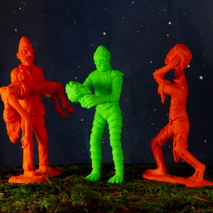 Pu&ograve; includere: Tre figure di giocattolo di plastica, una verde e due arancioni, in piedi su una superficie verde. La figura verde &egrave; un mostro con una texture di pelle squamosa. Una figura arancione porta una figura arancione pi&ugrave; piccola. L'altra figura arancione si tiene la testa tra le mani.
