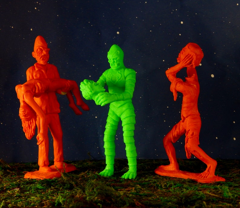 Pu&ograve; includere: Tre figure di giocattolo di plastica, una verde e due arancioni, in piedi su una superficie verde. La figura verde &egrave; un mostro con una texture di pelle squamosa. Una figura arancione porta una figura arancione pi&ugrave; piccola. L'altra figura arancione si tiene la testa tra le mani.