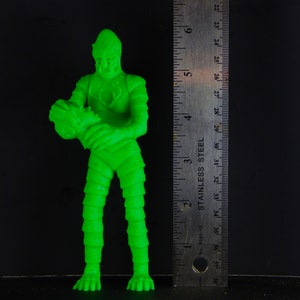 Pu&ograve; includere: Una action figure di plastica verde di una creatura con un corpo squamoso e testurizzato e una grande testa. La figura tiene una figura verde pi&ugrave; piccola nelle sue mani.
